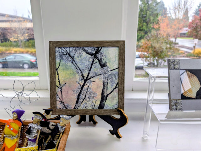 The image shows a framed artwork displayed on a windowsill. The artwork depicts dark tree branches with delicate leaves, set against a softly blended background of pastel colors - greens, yellows, and pinks.