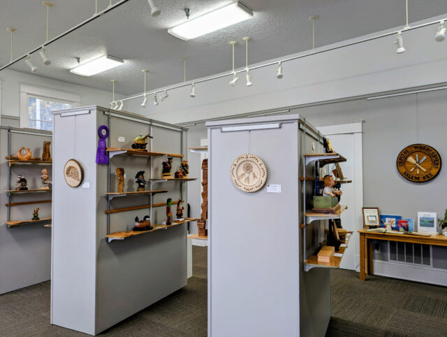 A well-lit art gallery space featuring woodcarvings. The room has several gray display panels with wooden shelves attached, each holding various carved wooden sculptures and reliefs: detailed figures, animals, people, and decorative plaques.